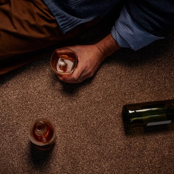 Man sitting on carpeted floor holding half-empty glass with bottle lying nearby.
