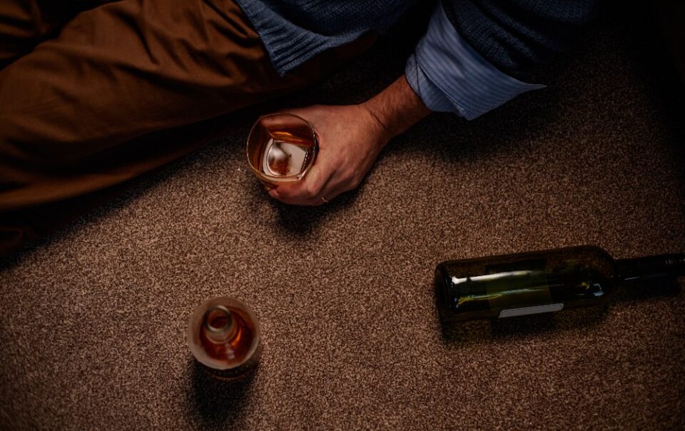 Man sitting on carpeted floor holding half-empty glass with bottle lying nearby.
