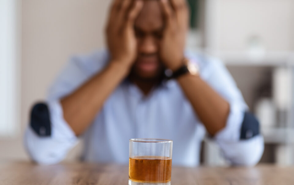 A man sitting his his hands on his head in front of a glass of alcohol wondering am i addicted to alcohol.