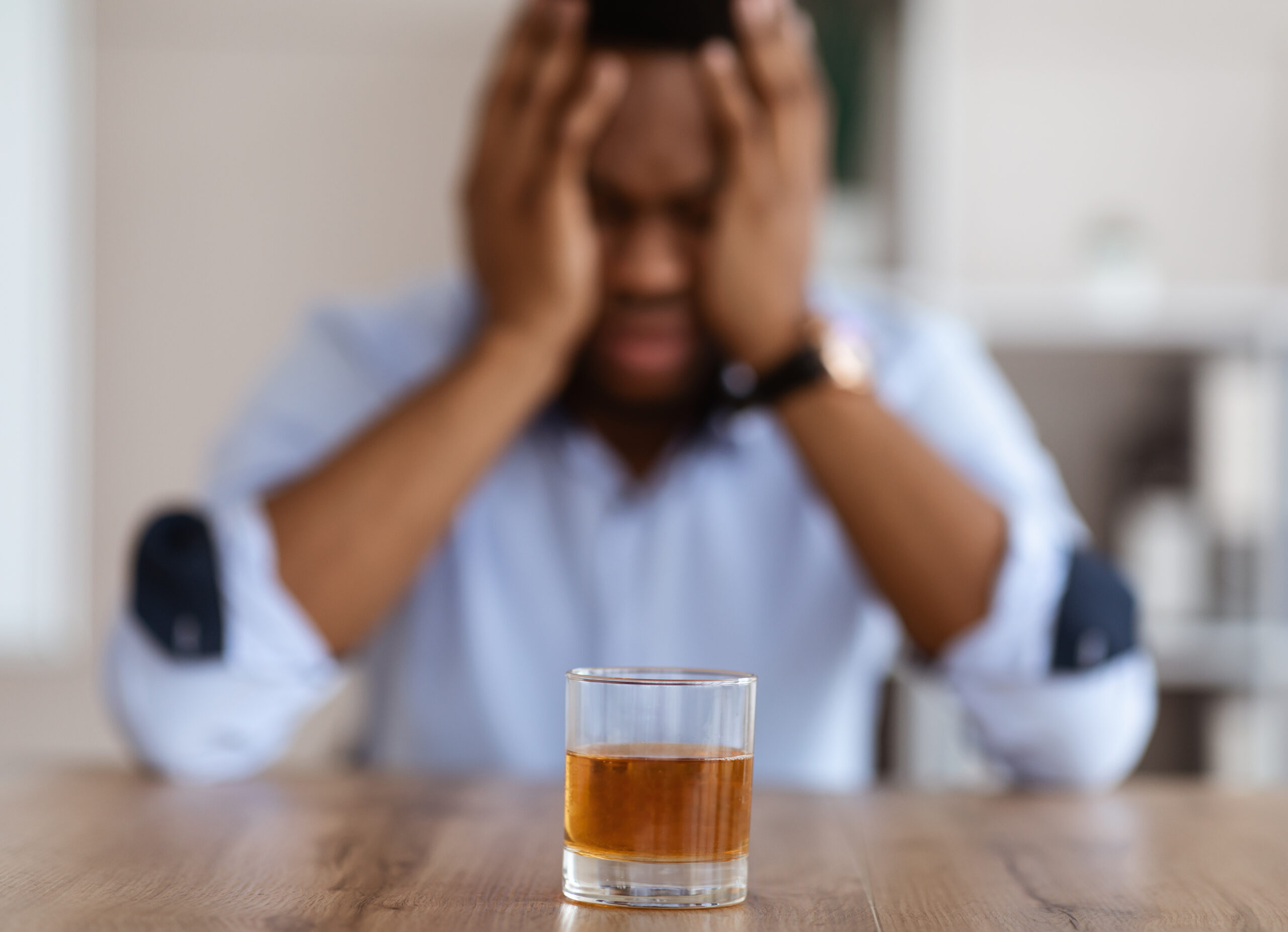 A man sitting his his hands on his head in front of a glass of alcohol wondering am i addicted to alcohol.